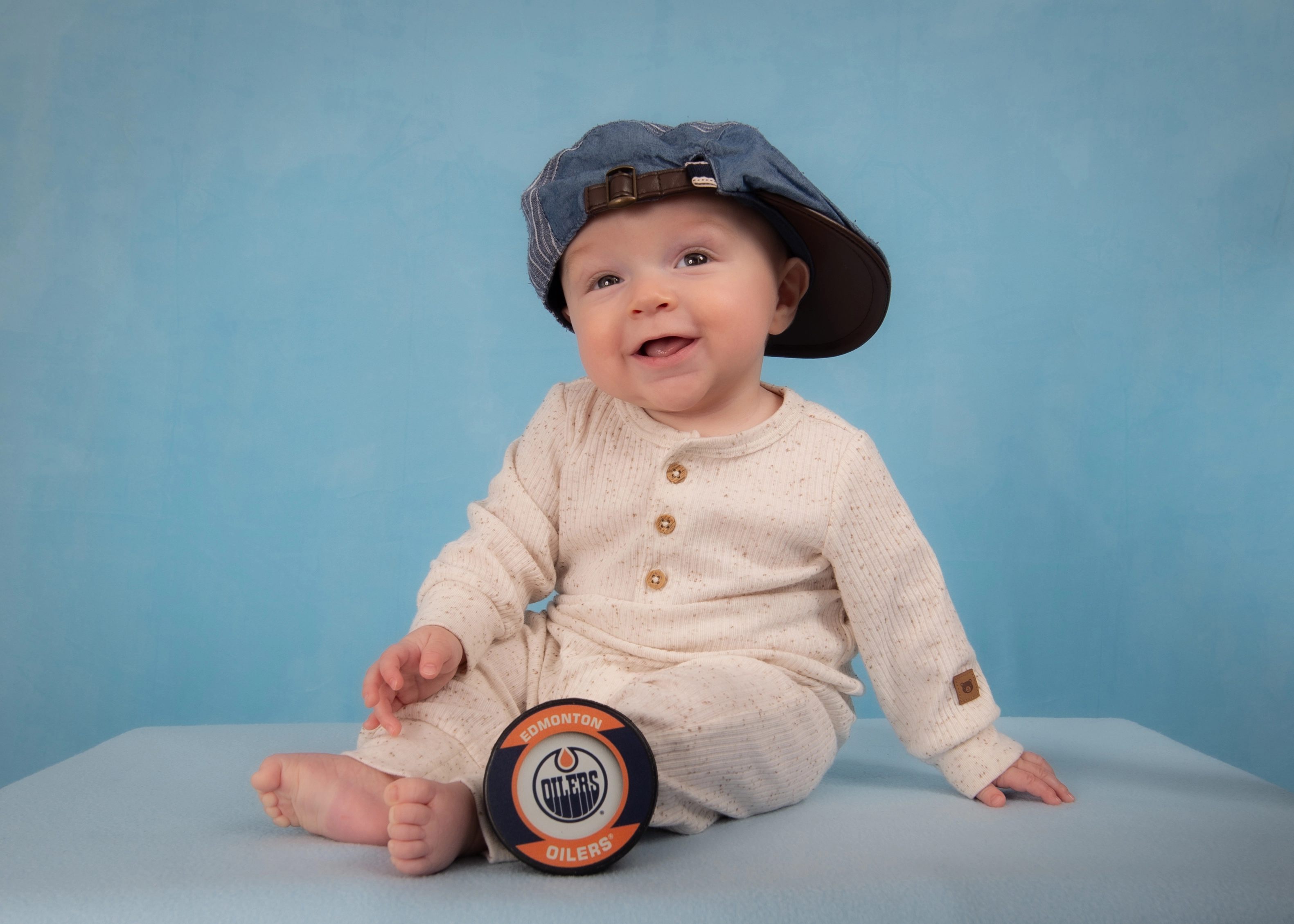 Smiling baby in denim cap with Edmonton Oilers puck on light blue studio backdrop — Edmonton baby milestone photo